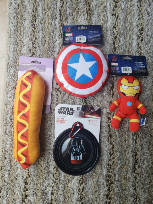 A collection of Marvel and Star Wars themed plush toys and accessories arranged on a textured beige carpet. The items include a hot dog plush, a Captain America shield plush, an Iron Man plush, a Darth Vader squeaker toy, and two packaged Avengers-themed items.