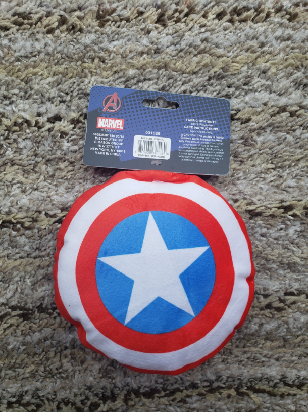 Captain America shield plush toy with its original retail packaging, placed on a textured beige carpet. The packaging features a blue header card with the Marvel logo.